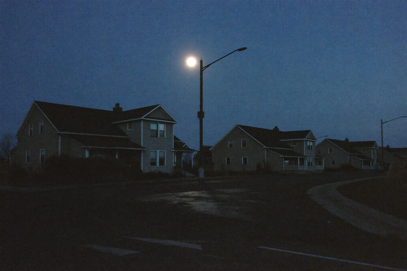 Darkened residential street during a nighttime power outage, every window black and the street light unlit, houses rim-lit by moonlight